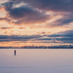 Una persona solitaria si trova su una vasta pianura innevata, osservando lo skyline di una città sotto un drammatico cielo al tramonto.