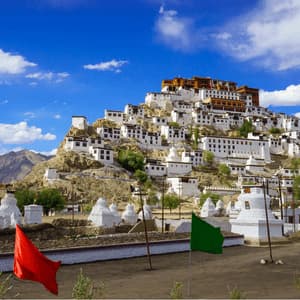 A white monastery complex built into a rocky hillside under a blue sky, with white stupas and colorful flags in the foreground.