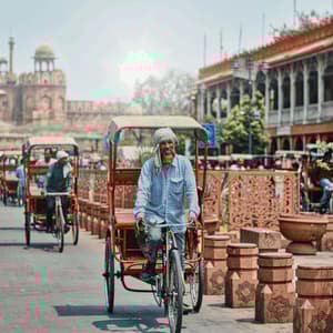 An elderly man with a turban rides a red cycle rickshaw on a busy street, with a historic fort visible in the background.