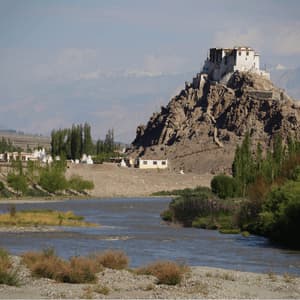 A white monastery sits on a rocky hill overlooking a river valley, with distant hazy mountains in the background.