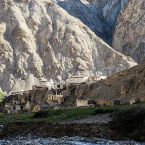 A small stone village with white-topped buildings is nestled into a grassy hillside at the base of a large, rugged mountain.
