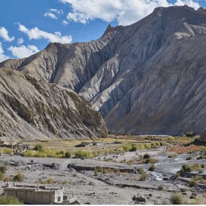A small village sits in a wide, arid valley at the base of large, rocky mountains under a partly cloudy blue sky.