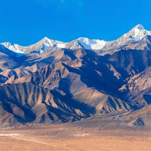 A rugged mountain range with snow-capped peaks stands against a clear blue sky, with a vast desert landscape in the foreground.