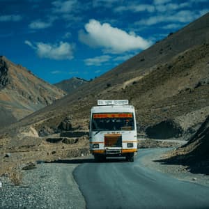 A white bus drives on a winding asphalt road through a rocky, mountainous landscape under a bright blue sky.