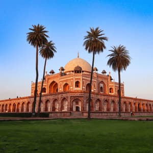 A red and white stone mausoleum with a central dome stands behind a green lawn, with four tall palm trees in front under a clear sky.