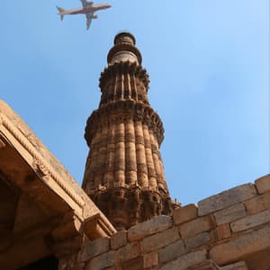 A low-angle view of an airplane flying over a tall, ornate sandstone tower against a clear blue sky, with ancient stone walls in the foreground.
