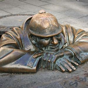 Una estatua de bronce de un hombre con sombrero emerge de una alcantarilla abierta en una acera urbana pavimentada, apoyando los brazos en el suelo.