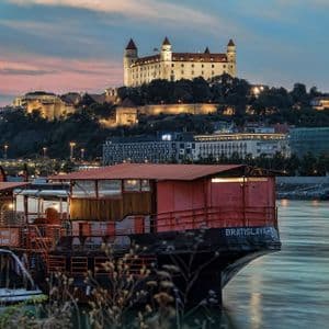 Un barco rojo está amarrado en un río con un castillo blanco iluminado en una colina al fondo al anochecer.