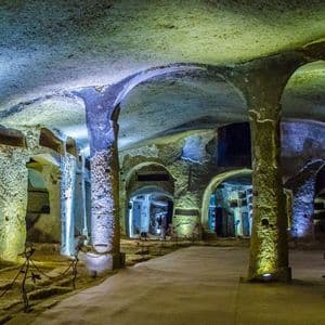 A large underground catacomb with rough-hewn stone pillars and arches, illuminated by blue and yellow lights.