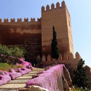 Una escalera de piedra bordeada por cascadas de flores moradas conduce a una gran fortaleza antigua con almenas contra un cielo azul claro.