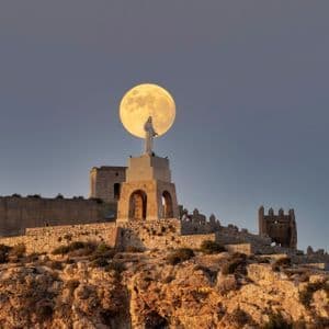 Una gran luna llena se alinea perfectamente detrás de una estatua sobre un pedestal en un castillo de piedra, sobre una colina rocosa al anochecer.