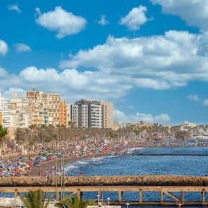 Una vista amplia de una concurrida playa urbana en un día soleado, con gente nadando y relajándose bajo sombrillas coloridas cerca de edificios y palmeras.