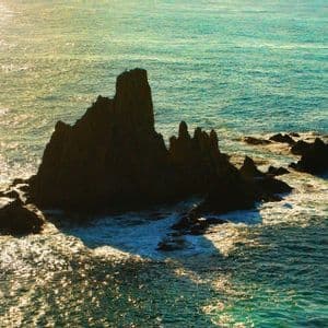 Un cúmulo de rocas oscuras y dentadas se eleva del mar turquesa, rodeado de agua brillante por el sol y espuma blanca.