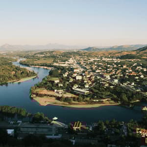 An aerial view of a river curving around a town, with a bridge, green hills, and distant mountains under a clear sky.