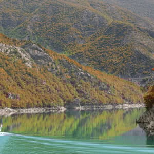 A tour boat carrying a WeRoad group trip navigates a calm lake, surrounded by steep mountains covered in autumn foliage.