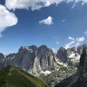 A green grassy ridge leads towards a jagged mountain range with patches of snow under a blue sky with white clouds.