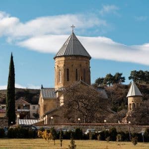 Un monastère en pierre avec un toit conique se dresse sur une pelouse herbeuse, entouré d'arbres, sous un ciel partiellement nuageux.