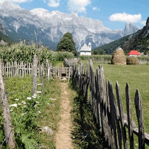 A dirt path along a wooden fence leads through a green valley with a church, cornfield, and tall mountains in the background.