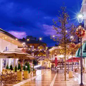 A wet pedestrian street lined with outdoor cafes is illuminated by string lights and decorated with Christmas wreaths at twilight.