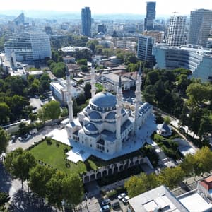 An aerial view of a large mosque with blue domes and four minarets, surrounded by green trees and a modern cityscape.