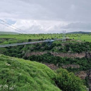 Un pont suspendu en verre avec une structure en forme de diamant au centre enjambe un canyon profond et verdoyant sous un ciel nuageux.