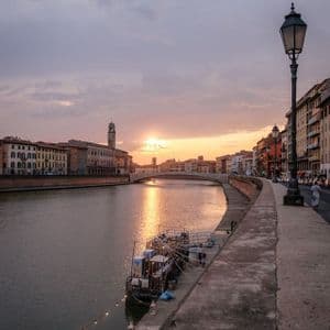 Ein breiter Fluss schlängelt sich bei Sonnenuntergang durch eine Stadt mit alten Gebäuden, das Sonnenlicht spiegelt sich glitzernd auf dem Wasser.