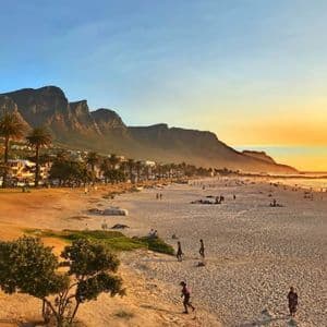 Un'ampia spiaggia sabbiosa con molte persone che si godono il tramonto, sullo sfondo di una grande catena montuosa e palme.