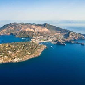 Una vista aerea di un'isola vulcanica con un grande cratere e una piccola città, circondata da un mare blu profondo sotto un cielo sereno.