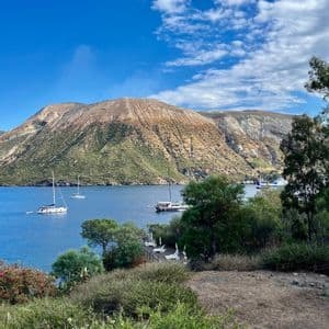 Una vista panoramica di una baia con diverse barche a vela, con una grande isola aspra sullo sfondo sotto un cielo azzurro con nuvole sparse.