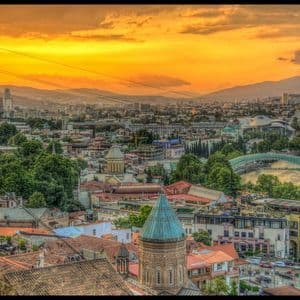Une vue en hauteur d'une ville avec des bâtiments historiques et modernes, un fleuve et des montagnes sous un coucher de soleil doré.