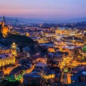 Une ville à flanc de colline avec des bâtiments historiques et une forteresse illuminée par des lumières chaudes au crépuscule, avec des montagnes en arrière-plan.
