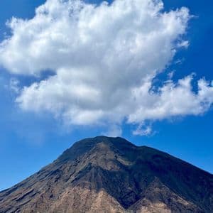 Un grande monte vulcanico sorge dal mare blu, la sua cima in ombra sotto una singola grande nuvola bianca in un cielo azzurro brillante.