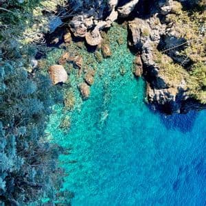 Una vista aerea di una costa rocciosa dove l'acqua limpida e turchese incontra la riva, rivelando rocce sotto la superficie.
