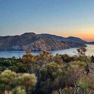 Due persone di un viaggio di gruppo WeRoad camminano su un sentiero collinare che domina una baia con isole vulcaniche al tramonto.