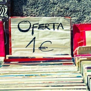 A handwritten sign reading 'Oferta 1€' sits atop stacks of secondhand vinyl records at an outdoor market stall.