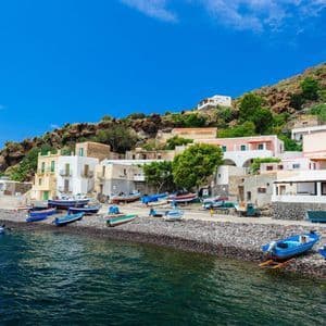 Piccole barche sono ormeggiate su una spiaggia di ciottoli di fronte a un villaggio incastonato su un fianco di collina roccioso, sotto un cielo azzurro.
