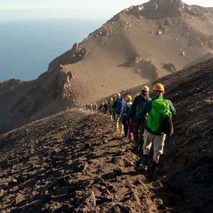 Un gruppo WeRoad con i caschi fa trekking in fila indiana su un ripido sentiero di montagna vulcanico a picco sull'oceano.
