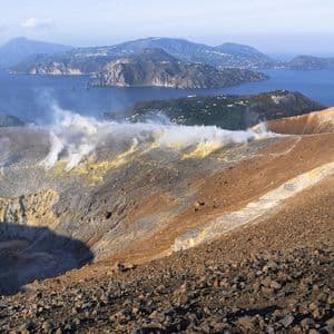 Il vapore sale dai pendii sulfurei di un cratere vulcanico, con vista su isole oltre il mare sullo sfondo.