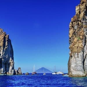 Diverse barche sull'acqua blu passano tra due grandi formazioni rocciose, con un vulcano visibile in lontananza sotto un cielo sereno.