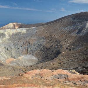 Una vista panoramica di un grande cratere vulcanico con vapore che sale dal suo bordo, con il mare visibile sullo sfondo sotto un cielo blu.