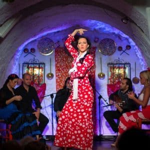 Una mujer con un vestido de lunares rojos y blancos baila flamenco en un escenario en un local tipo cueva, acompañada por músicos.