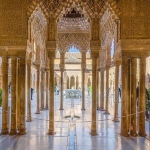 Columnas y arcos esbeltos y ornamentados rodean un patio de palacio soleado con una fuente central y un estrecho canal de agua.