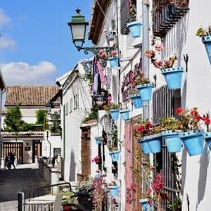 Una calle estrecha y soleada de adoquines con edificios encalados, uno de los cuales está decorado con muchas macetas azules.