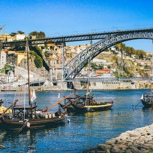 Barche tradizionali in legno galleggiano su un fiume con un grande ponte ad arco in metallo e una città collinare sullo sfondo.