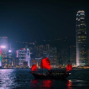 Una tradizionale giunca con vele rosse illuminate galleggia sull'acqua di notte davanti a uno skyline cittadino splendidamente illuminato.