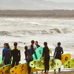 Un groupe WeRoad, en combinaisons de surf et avec des planches, se tient sur une plage, observant les vagues de l'océan avant une leçon de surf.