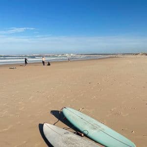 Two surfboards rest on a sandy beach with people by the ocean in the background under a clear blue sky.