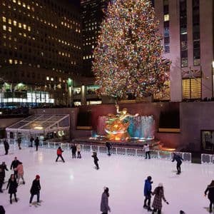 Des patineurs glissent sur une patinoire extérieure la nuit, devant un grand sapin de Noël illuminé et une statue dorée, en pleine ville.