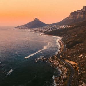 Una vista aerea di una strada tortuosa lungo una costa rocciosa, con montagne e una città in lontananza sotto un cielo arancione al tramonto.