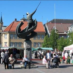 La gente se reúne en un mercado al aire libre en una plaza, centrado alrededor de una gran estatua de un hombre en un barco.